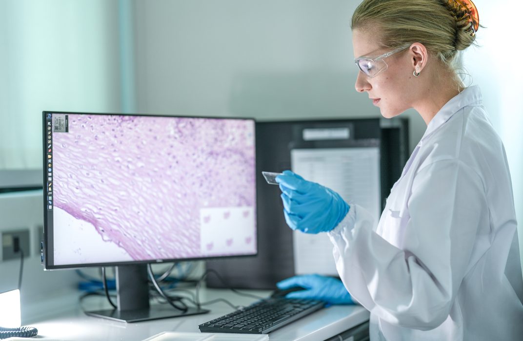 A researcher with a tablet and a laptop at a desk in a laboratory office.