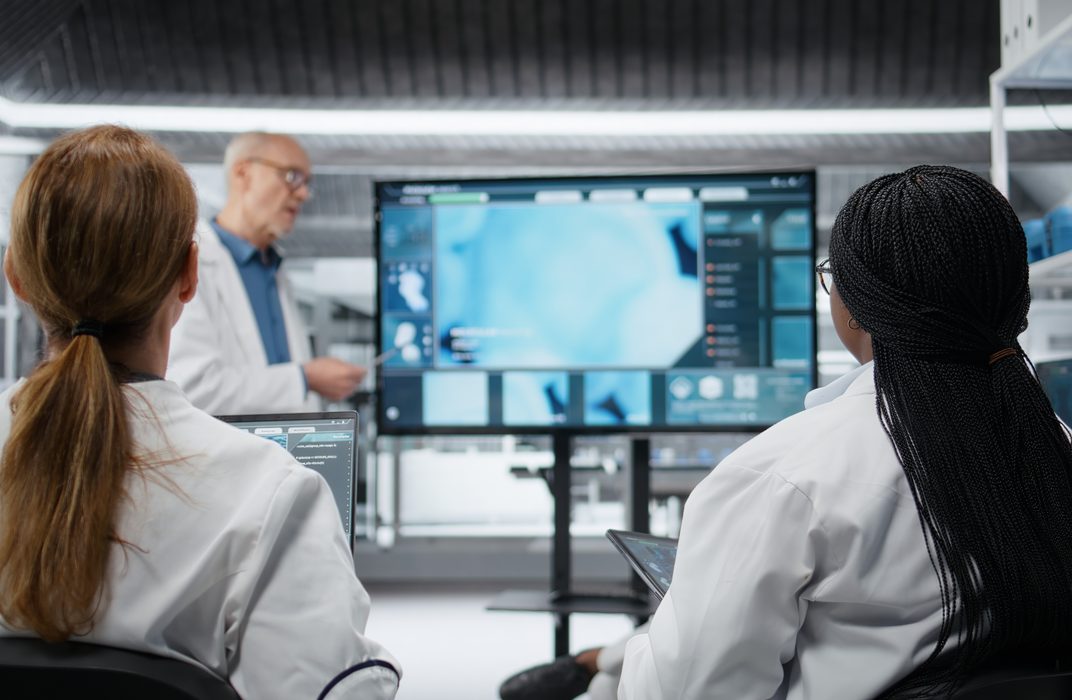 Two scientists in lab coats discuss a stained tissue image on a monitor.